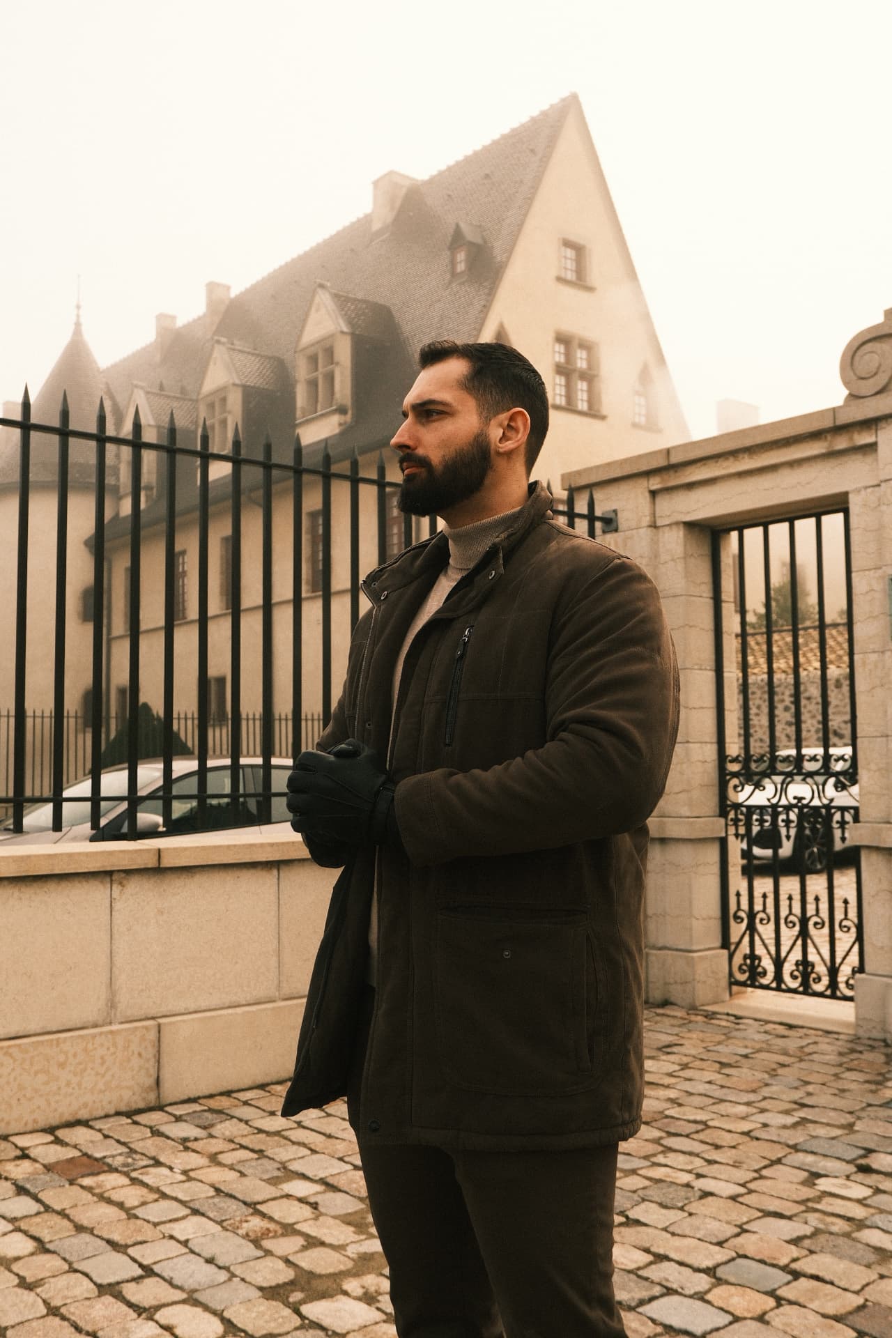 Homme debout sur un pavement, devant le portail en fer forgé d'une maison de maître enveloppée de brume