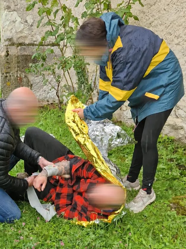 Exercice de secourisme en extérieur : deux personnes soignent une personne allongée sur l’herbe, bande de pansement et couverture de survie, visages non identifiables.